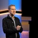 Geoff Keighley clasps his hands on stage as a zombie walks towards him in the background.