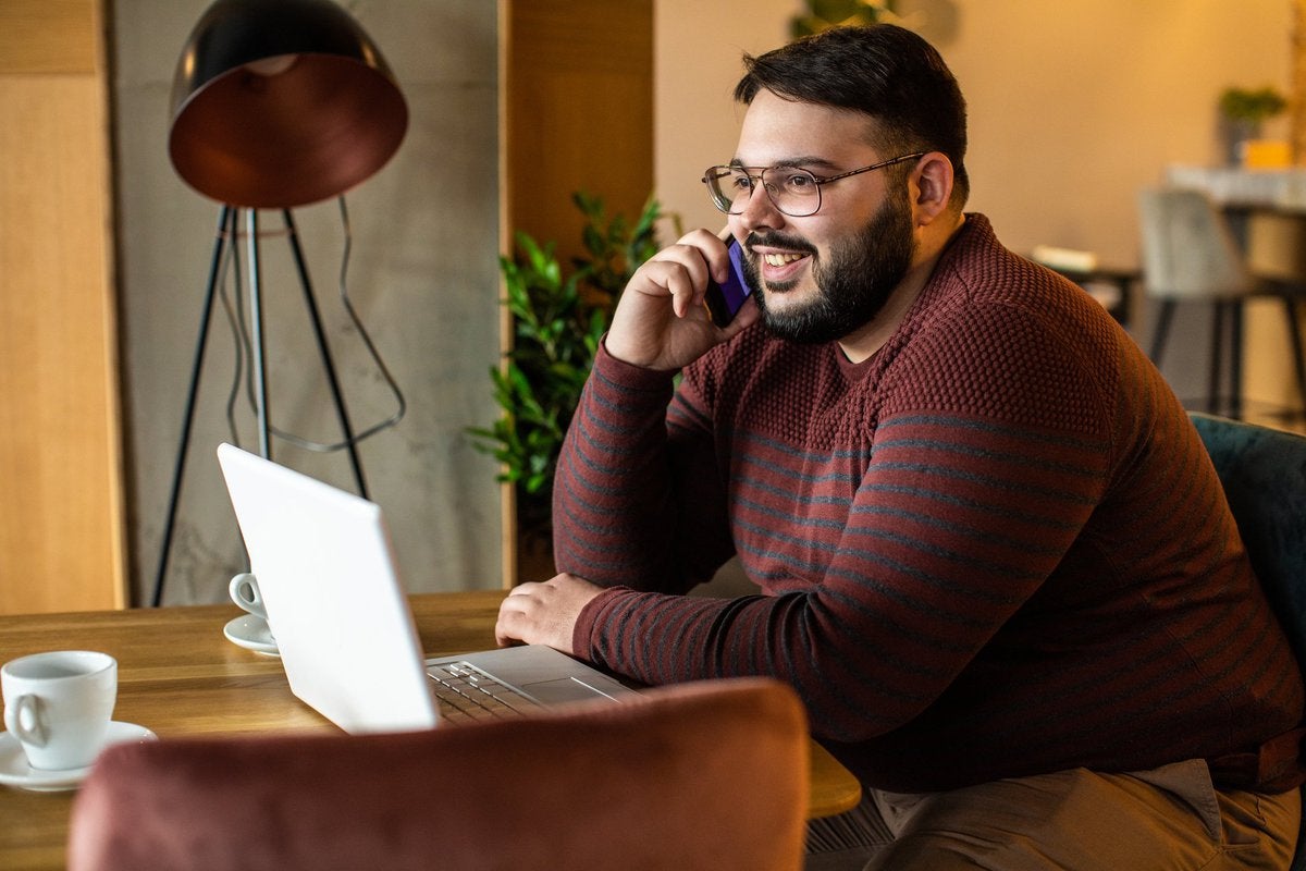 A man talks on the phone while sitting in front of his laptop in a coffee shop.