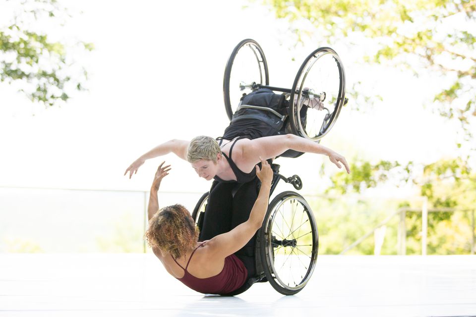 Kinetic Light's Alice Sheppard and Laurel Lawson will perform at the 2022 Jacob's Pillow Gala. Here, Alice lifts Laurel up from below while stretching her arms and torso up in a hug. Laurel hugs back, arms open and bent, wheels turning.