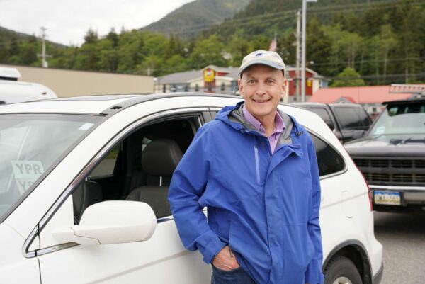 A man in a blue windbreaker leans against a white car